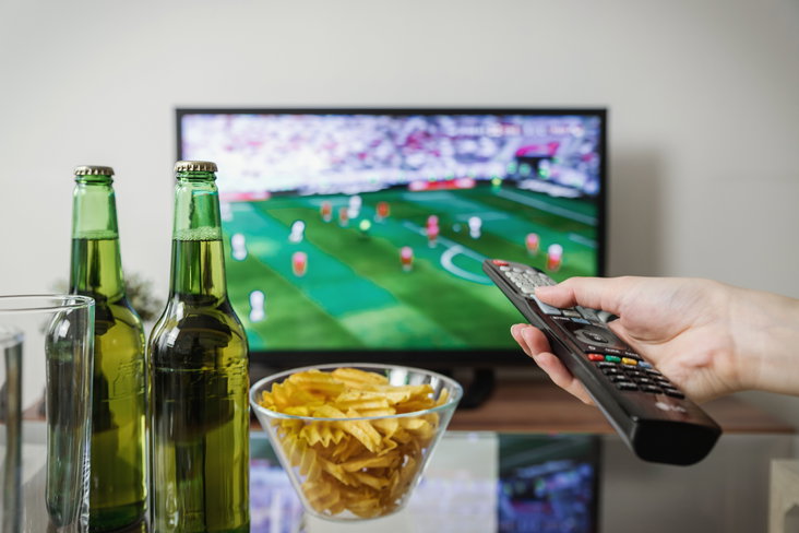 Iemand houdt een afstandsbediening vast terwijl hij naar voetbal op tv kijkt, met bier en snacks op tafel.